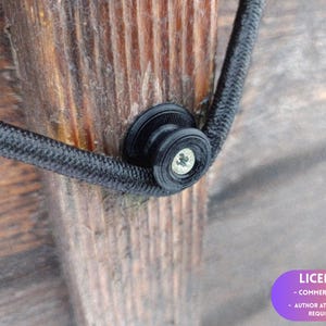 May include: A black circular plastic cord guide with a central silver-coloured screw, mounted onto a vertical wooden surface. A black braided cord is wrapped around the groove of the guide, which features a layered, ridged texture.