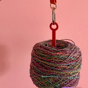 Pode incluir: Uma bola de lã colorida com uma ferramenta vermelha em forma de chave presa a uma correia de couro vermelha. A lã é uma mistura de fios verdes, roxos e amarelos. O fundo é de cor rosa lisa.