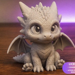 Puede incluir: Figura de dragón gris claro, sentado, con grandes ojos expresivos y pequeñas alas. El dragón tiene una apariencia texturizada y escamosa. La figura tiene un diseño lindo y de dibujos animados, sobre una superficie de madera.