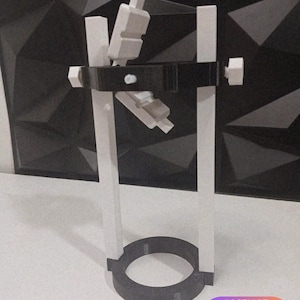Pode incluir: Um suporte vertical com dois postes paralelos brancos conectados por uma base circular preta e uma barra transversal preta. A barra transversal inclui botões de ajuste brancos e um componente articulado central com dois blocos.