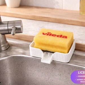 May include: A yellow Vileda sponge rests in a white plastic holder on a stainless steel sink. The sponge has red lettering. A chrome faucet is visible on the left. The background includes a wooden shelf and small plants.