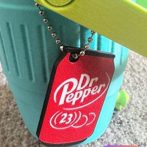 May include: A red Dr Pepper tag with white text and the number 23, attached to a teal bucket with a green handle. The tag is hanging from a silver ball chain. The image includes the text "@CHACHACHA".