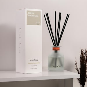 May include: A white box and a frosted glass reed diffuser with black reeds. The box reads "Amber Sandalwood" and "Nesti Casa". The diffuser has an orange band and the same text as the box. The product is on a white shelf.