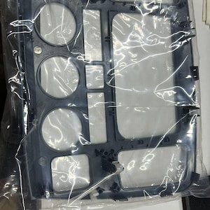 Puede incluir: Un componente nuevo de salpicadero de coche de plástico gris oscuro, aún en su embalaje original. El componente tiene varias aberturas para controles y pantallas, incluyendo recortes circulares y rectangulares. El plástico parece liso y mate.