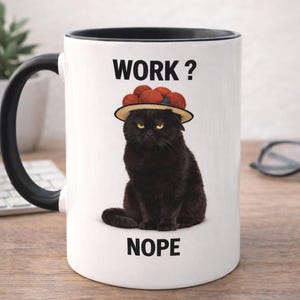 Könnte beinhalten: Weißer Keramikbecher mit schwarzem Henkel und Rand. Der Becher zeigt eine schwarze Katze mit Hut und roten Pompons. Der Text "WORK?" und "NOPE" sind auf dem Becher aufgedruckt.