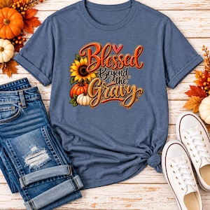 May include: A heather blue short-sleeve t-shirt featuring a graphic print with the text Blessed Beyond the Gravy in orange and brown script. The design includes a yellow sunflower, an orange pumpkin, and a small white pumpkin.