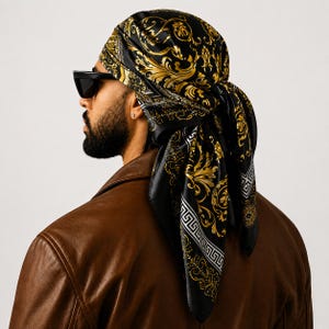 May include: A square silk headscarf featuring a black background with intricate gold baroque scrollwork patterns and a white Greek key border design. The scarf is tied around the head, displaying a glossy finish and detailed ornamental motifs.