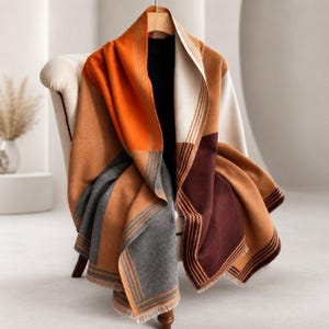 May include: A large, rectangular wrap or shawl featuring a color-blocked design in shades of orange, tan, dark brown, and grey. The fabric displays a series of thin, parallel vertical stripes near the edges and includes a fringed hem detail.