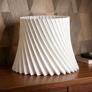 May include: A white, sculptural lampshade with a spiral design. The shade has a wide, open top and a flared base. The design features vertical, angled lines that create a textured surface. The lampshade is placed on a dark wooden surface.