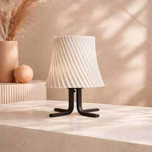 May include: A modern table lamp with a cream-colored, pleated lampshade. The lampshade sits atop a black, stylized base. The lamp is on a light-colored surface, with a vase and decorative sphere in the background.