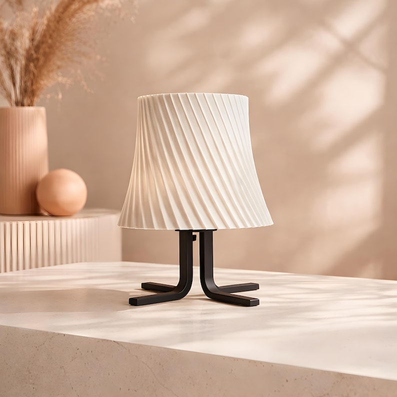 May include: A modern table lamp with a cream-colored, pleated lampshade. The lampshade sits atop a black, stylized base. The lamp is on a light-colored surface, with a vase and decorative sphere in the background.