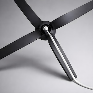 May include: A modern black ceiling fan with four blades. The fan's blades are angled and extend from a central hub. A white cord runs along the edge of one blade. The fan is set against a gray background.