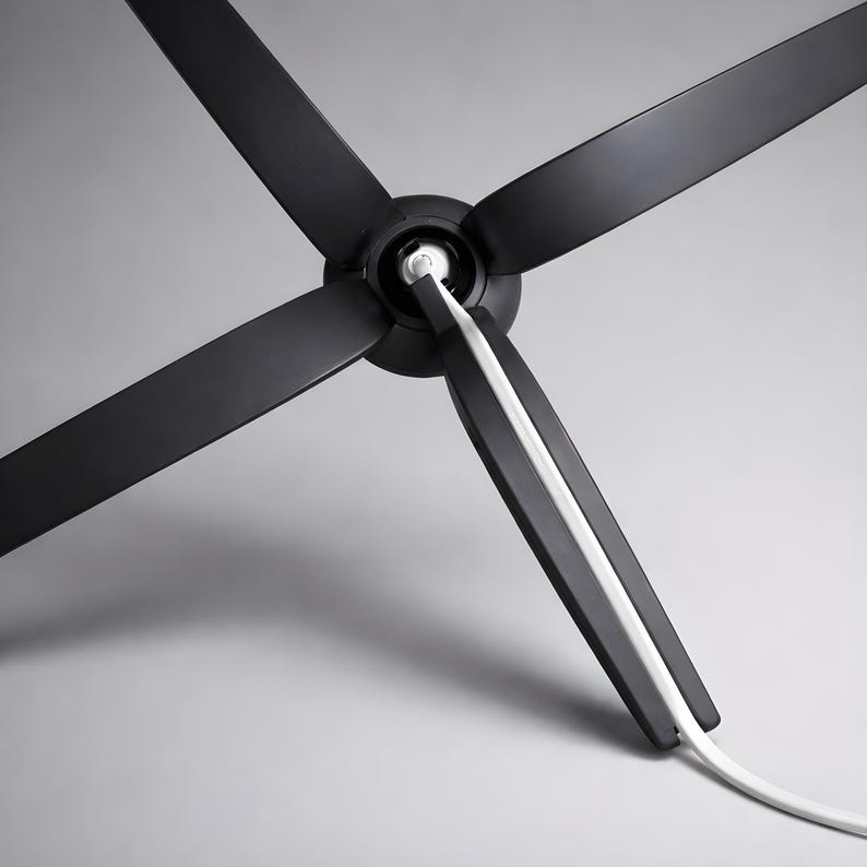 May include: A modern black ceiling fan with four blades. The fan's blades are angled and extend from a central hub. A white cord runs along the edge of one blade. The fan is set against a gray background.