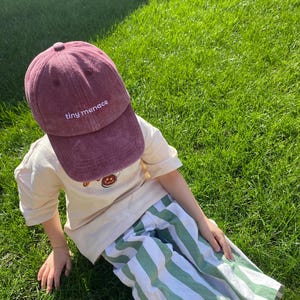 May include: A burgundy baseball cap with the text "tiny menace" embroidered on the front. The cap is worn by a person wearing a cream-coloured t-shirt and green and white striped trousers. The person is sitting on green grass.