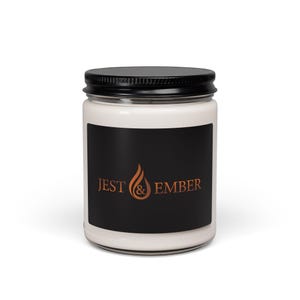 Op de afbeelding: Een witte kaars in een heldere glazen pot met een zwart deksel. Een zwart etiket op de voorkant toont de tekst "JEST & EMBER" in een koperkleurig lettertype, met een vlammenafbeelding.