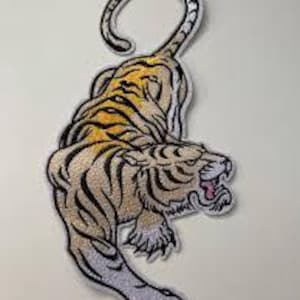 May include: Embroidered tiger patch featuring a detailed design. The tiger is shown in tan, black, and yellow, with its mouth open, showing pink. The patch has a white border and is suitable for application on clothing or accessories.