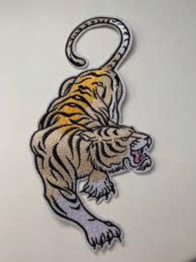 May include: Embroidered tiger patch featuring a detailed design. The tiger is shown in tan, black, and yellow, with its mouth open, showing pink. The patch has a white border and is suitable for application on clothing or accessories.