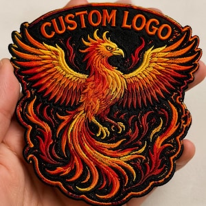 May include: Embroidered patch featuring a phoenix with outstretched wings, rendered in shades of red, orange, and yellow against a black background. The patch has the text "CUSTOM LOGO" in orange. The phoenix is surrounded by flames.