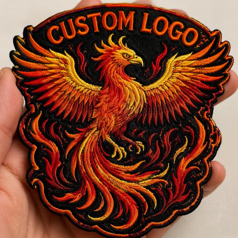 May include: Embroidered patch featuring a phoenix with outstretched wings, rendered in shades of red, orange, and yellow against a black background. The patch has the text "CUSTOM LOGO" in orange. The phoenix is surrounded by flames.