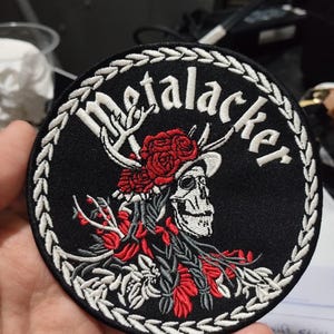 Könnte beinhalten: Runder schwarzer Aufnäher mit dem Wort "Metalacker" in Weiß, mit einem Totenkopf mit Geweih und roten Rosen. Das Design ist von einem weißen, gestickten Rand umgeben. Ein einzigartiger Aufnäher für Jacken, Taschen oder andere Kleidungsstücke.