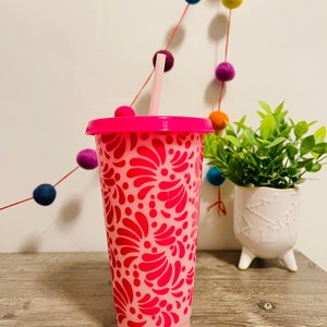 May include: A pink reusable tumbler with a pink and white floral design. The tumbler has a lid and a straw.