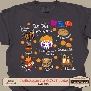 Peut inclure: T-shirt gris foncé avec un motif du Dia de los Muertos. Le motif comprend un papillon, une tête de mort, du pain, des fleurs et d'autres éléments culturels. Le texte comprend "tis the season" et des mots espagnols.