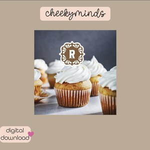 May include: A close-up of cupcakes with white frosting and a brown decorative topper with the letter "R" in the center. The cupcakes are on a gray surface.