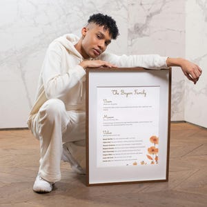 May include: A framed print displaying the text "The Bryan Family". The print includes a list of values, mission, and vision statements, with orange floral accents. The frame is a dark brown colour.