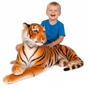 May include: A large plush tiger toy with orange and black stripes, lying on a white surface. The tiger has a detailed face with realistic features. A young child in a blue top is sitting on the tiger, smiling.