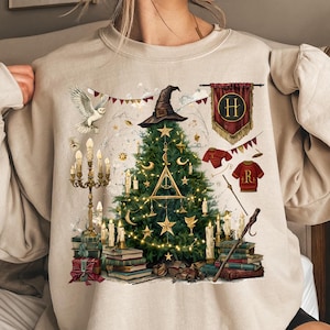 May include: A beige crewneck sweatshirt featuring a graphic print of a Christmas tree decorated with stars, moons, and a Deathly Hallows symbol. The design includes a witch hat, a white owl, a candelabra, stacks of books, and a red banner with an H.