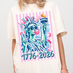 Może przedstawiać: Kremowy t-shirt z kolorową ilustracją Statuy Wolności w okularach przeciwsłonecznych i dmuchającej bańkę z gumy do żucia. Grafika zawiera niebieski tekst "1776-2026". Tło ma różowe i białe pionowe paski oraz abstrakcyjne pociągnięcia pędzla.