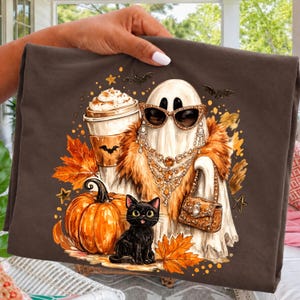 Könnte beinhalten: Ein dunkelbraunes T-Shirt mit einer Grafik eines Geistes, der eine Sonnenbrille, einen Pelzkragen und Schmuck trägt und eine Handtasche hält. Der Geist wird von einer schwarzen Katze, einem Kürbis, einem Kaffeebecher und Herbstblättern begleitet.