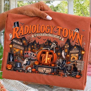 May include: A burnt orange t-shirt featuring a graphic of a spooky town with skeletons, pumpkins, and medical imaging equipment. The text reads RADIOLOGY TOWN WHERE WE SEE WHAT OTHERS MISS and EST. 1998, with various X-ray and MRI themed illustrations.