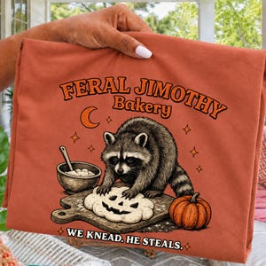 Könnte beinhalten: Ein rostorangefarbenes T-Shirt mit einer Grafik eines Waschbären, der Teig auf einem Holzbrett neben einer Schüssel und einem Kürbis knetet. Der Text lautet FERAL JIMOTHY BAKERY und WE KNEAD, HE STEALS, ergänzt durch einen Halbmond und Sternsymbole.