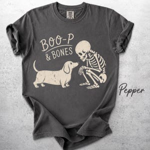May include: A charcoal gray short-sleeve t-shirt featuring a cream-colored graphic of a dachshund dog facing a human skeleton. The text above the dog reads BOO-P & BONES, and the word Pepper is printed in cursive script on the lower right side.