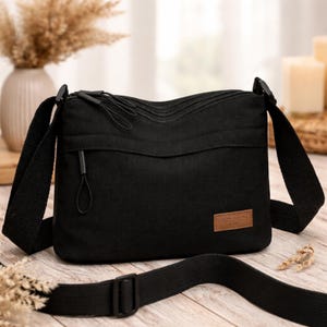 May include: A black canvas crossbody bag featuring multiple zippered compartments with black cord pulls. The bag includes an adjustable black fabric shoulder strap and a rectangular brown leather-like patch on the front with the text SCSIDI.