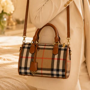 Könnte beinhalten: Eine zylinderförmige Handtasche mit einem Karomuster in Beige, Schwarz, Weiß und Rot. Die Tasche verfügt über braune Ledergriffe, einen passenden abnehmbaren Schulterriemen, goldfarbene Metallbeschläge und einen kleinen Lederanhänger.