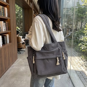 Puede incluir: Un bolso tote de pana gris con un bolsillo frontal grande y dos bolsillos laterales. El bolso tiene una correa para el hombro y borlas decorativas. El bolso lo lleva una persona.