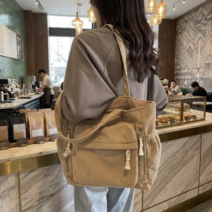 May include: A beige corduroy tote bag featuring a large front pocket with a flap closure, two side pockets with vertical zippers, and white knotted cord zipper pulls. The bag has a long shoulder strap and a textured, ribbed fabric construction.