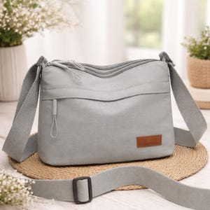 May include: A pale grey crossbody bag with an adjustable strap. The bag features a front zipped pocket and a brown rectangular brand patch. The bag is made from a durable material and is suitable for everyday use.