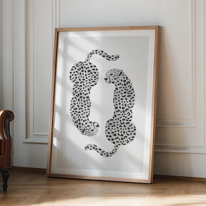 May include: A framed art print showing two stylized cheetahs in black and white. The cheetahs have spots and are arranged in a mirrored pose against a white background. The artwork is encased in a light brown wooden frame.