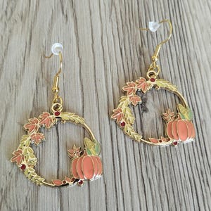 Può includere: Un paio di orecchini pendenti color oro con un design a corona circolare. Ogni orecchino è decorato con foglie autunnali in smalto nei toni dell'arancione, giallo e verde, accentuato da una piccola zucca arancione e minuscole bacche rosse.