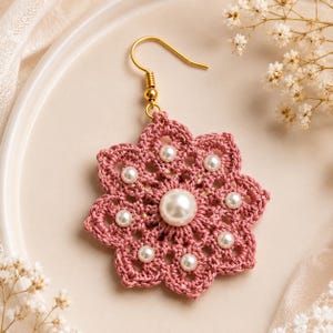 May include: A handcrafted, dusty rose crochet earring with a floral design. The earring features a gold-colored hook and is adorned with faux pearls. The crochet pattern creates an open, airy design, with a large pearl in the center and smaller pearls around it.