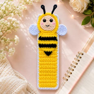 May include: A rectangular crochet bookmark featuring a bee character with a yellow and black striped body, a beige face with black eyes, a small smile, pink cheeks, and two black antennae. The bee has light blue wings on each side of its head.