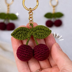 Puede incluir: Un llavero de cereza hecho a mano con ganchillo. El llavero presenta dos cerezas de color rojo oscuro con hojas verdes y un llavero dorado. Las cerezas se sostienen en una mano, con otros dos llaveros en el fondo.