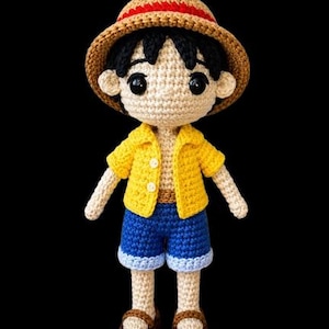 May include: A crocheted doll depicting a character in a straw hat with a red band, a yellow shirt, blue shorts, and brown sandals. The doll has black hair and large black eyes. The background is black.