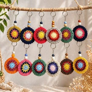 Può includere: Una collezione di portachiavi all'uncinetto fatti a mano in vari colori. Ogni portachiavi presenta un design circolare con un motivo a stella centrale e un accento di perline. Sono appesi a un ramo di legno.