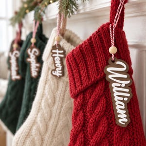 May include: A bright red knitted Christmas stocking featuring a thick cable knit pattern and a folded cuff. A wooden name tag with the white script text William is attached to the stocking with a red and white striped string and a small wooden bead.