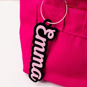May include: A pink luggage tag with the name "Emma" in a cursive font, outlined in black. The tag is attached to a bright pink tote bag with a white cord.