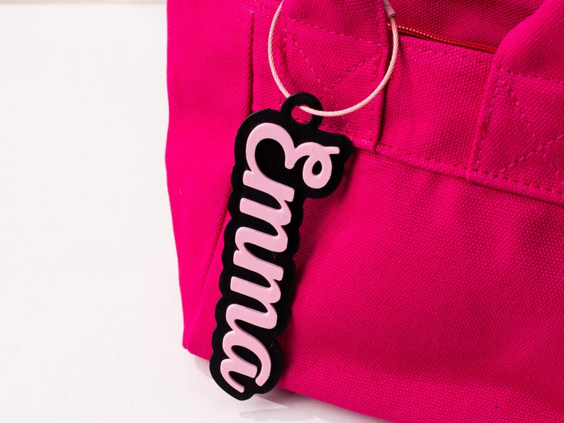 May include: A pink luggage tag with the name "Emma" in a cursive font, outlined in black. The tag is attached to a bright pink tote bag with a white cord.